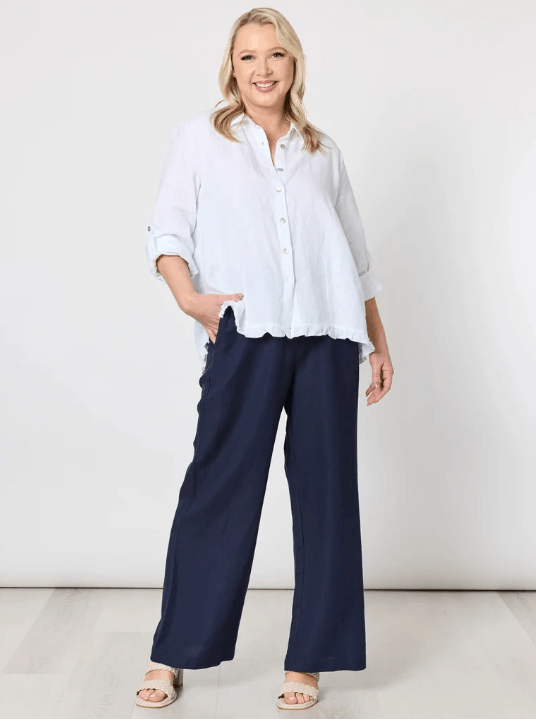 Gordon Smith woman  wearing navy wide-leg pants with a white button-up shirt and white espadrille wedge sandals, standing against a light gray wall and pale wood floor.