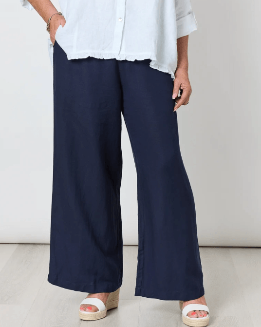Gordon Smith woman  wearing navy wide-leg pants with a white button-up shirt and white espadrille wedge sandals, standing against a light gray wall and pale wood floor.