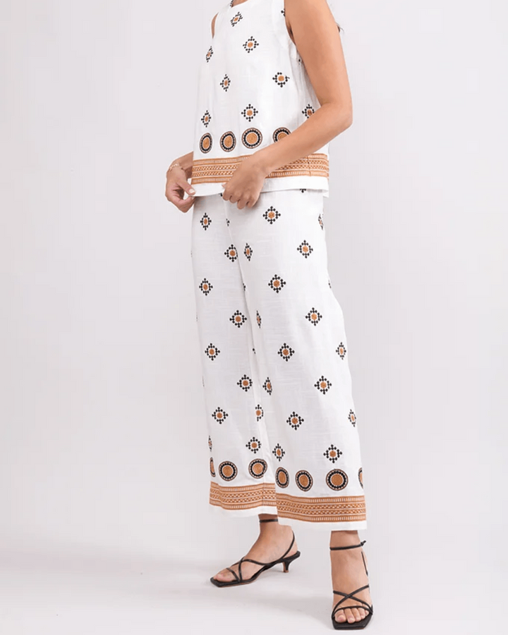Foil woman modeling a sleeveless white two-piece set with brown geometric patterns and tan striped trim, paired with black strappy heels, against a light gray studio background.