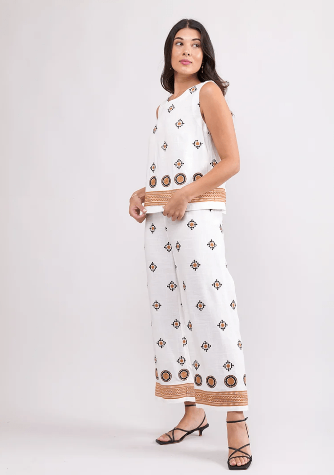 Foil woman modeling a sleeveless white two-piece set with brown geometric patterns and tan striped trim, paired with black strappy heels, against a light gray studio background.