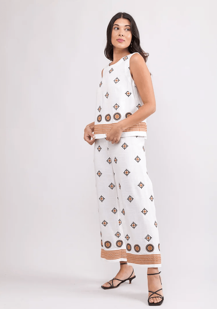 Foil woman modeling a sleeveless white two-piece set with brown geometric patterns and tan striped trim, paired with black strappy heels, against a light gray studio background.