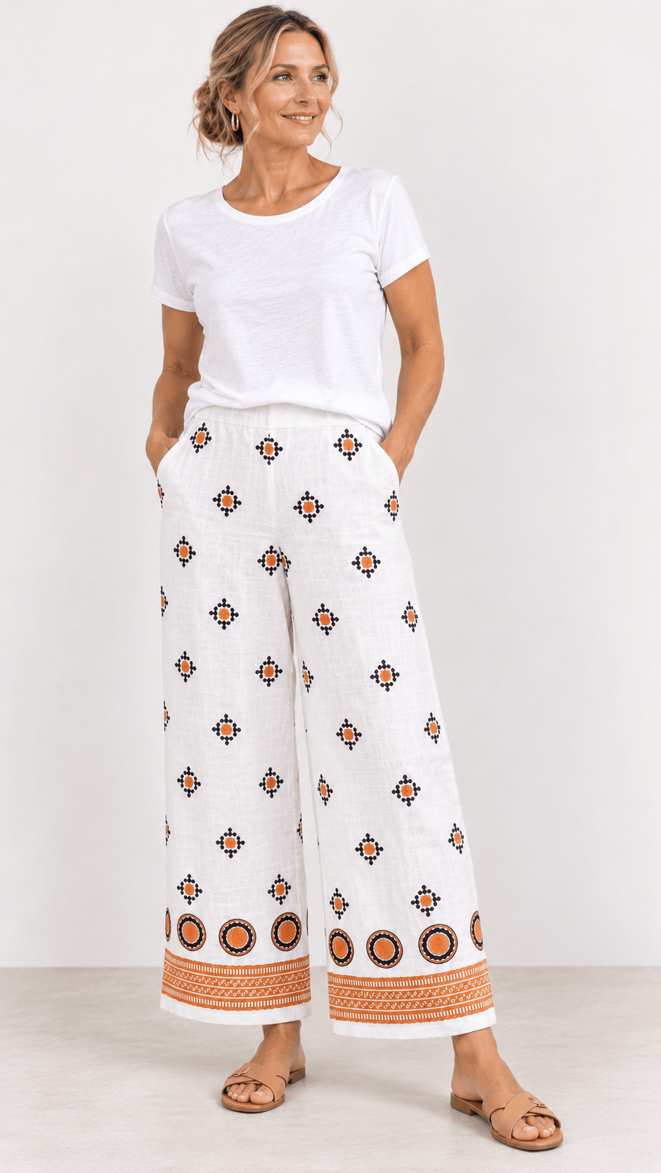 Foil woman modeling a sleeveless white two-piece set with brown geometric patterns and tan striped trim, paired with black strappy heels, against a light gray studio background.