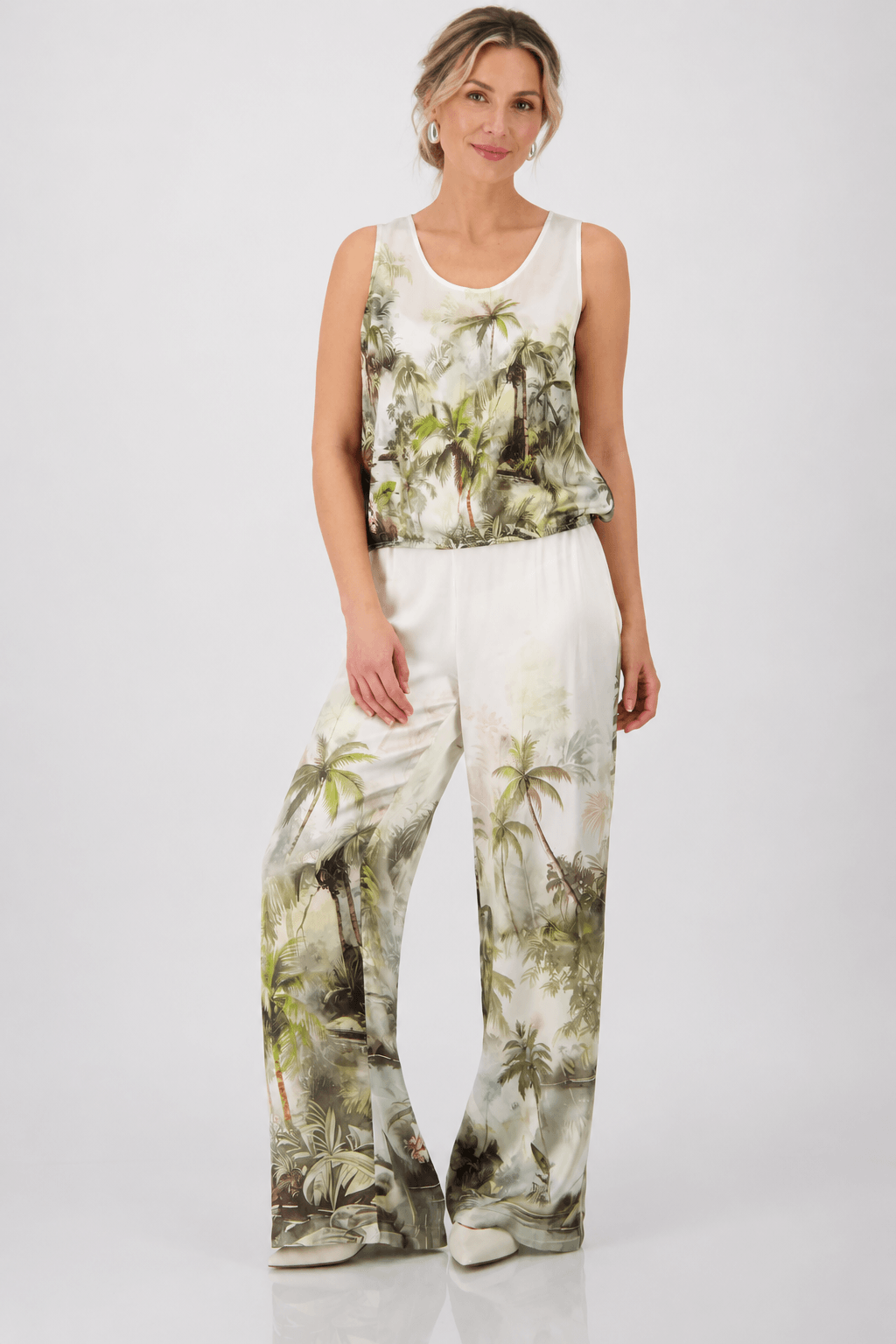 Monari modeling a sleeveless white tank top with a green and brown palm tree tropical print, paired with matching white pants, against a light gray studio background.