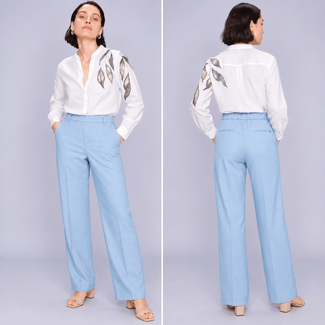 Mos Mosh woman modeling a white long-sleeve blouse with metallic gold leaf embroidery on the shoulders, paired with light blue wide-leg trousers and beige sandals, against a solid light gray studio background.