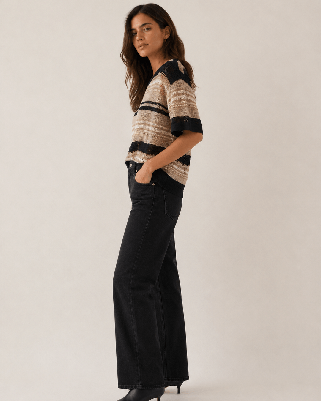 LD&amp;Co modeling wearing a short-sleeve knit sweater with horizontal beige, brown, and black stripes, paired with black jeans, against a plain light beige studio background.