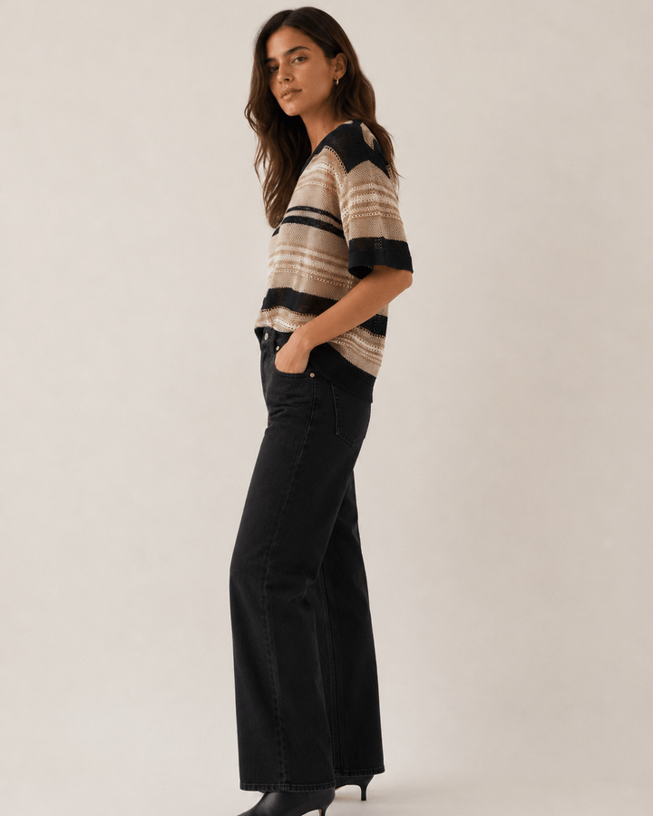 LD&amp;Co modeling wearing a short-sleeve knit sweater with horizontal beige, brown, and black stripes, paired with black jeans, against a plain light beige studio background.
