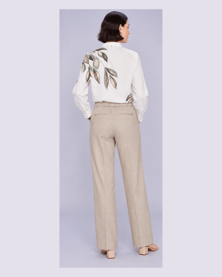Mos Mosh woman modelling from the back wearing a white long-sleeve blouse with a large leaf print on the back and beige wide-leg trousers, standing against a light gray studio background.