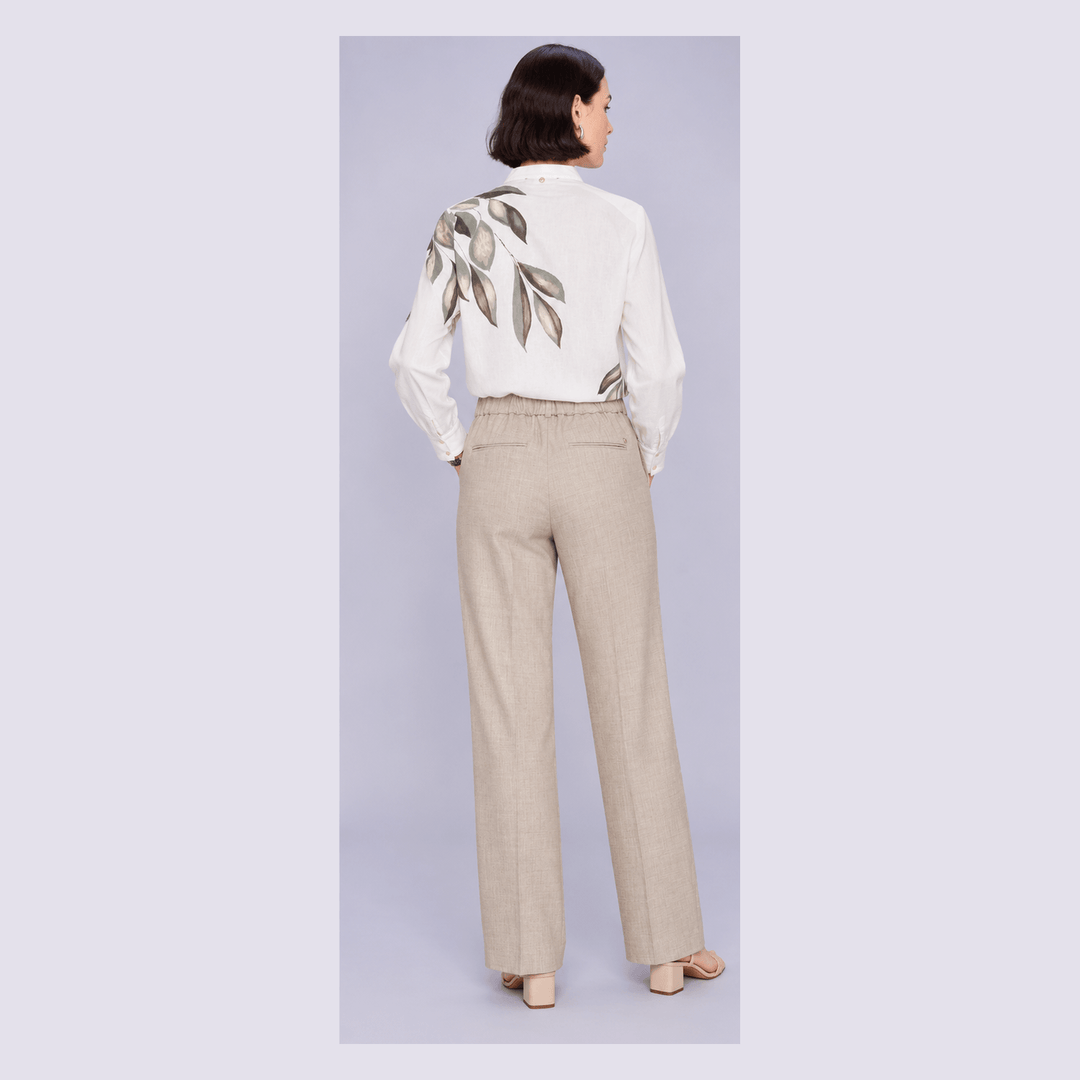 Mos Mosh modelling  from the back wearing a white long-sleeve blouse with a large leaf print on the back and beige wide-leg trousers, standing against a light gray studio background.