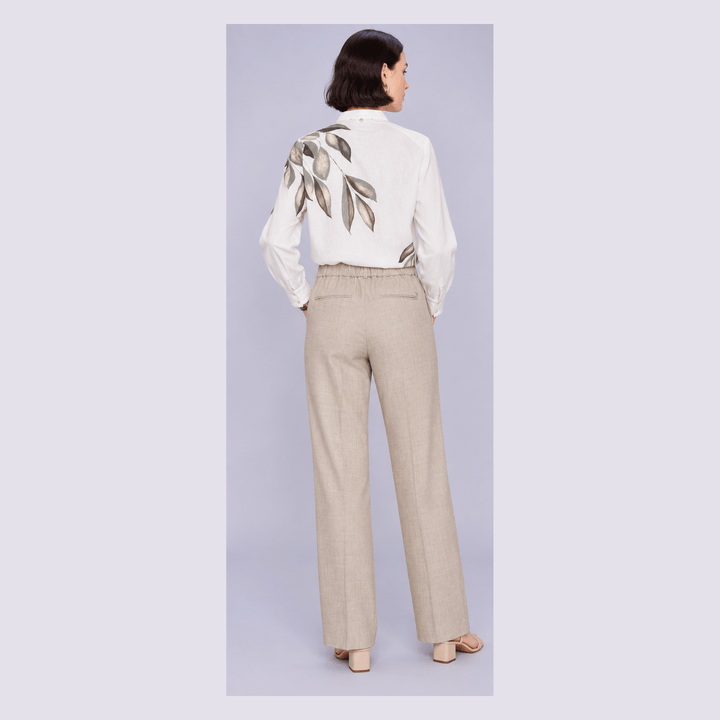 Mos Mosh modelling  from the back wearing a white long-sleeve blouse with a large leaf print on the back and beige wide-leg trousers, standing against a light gray studio background.
